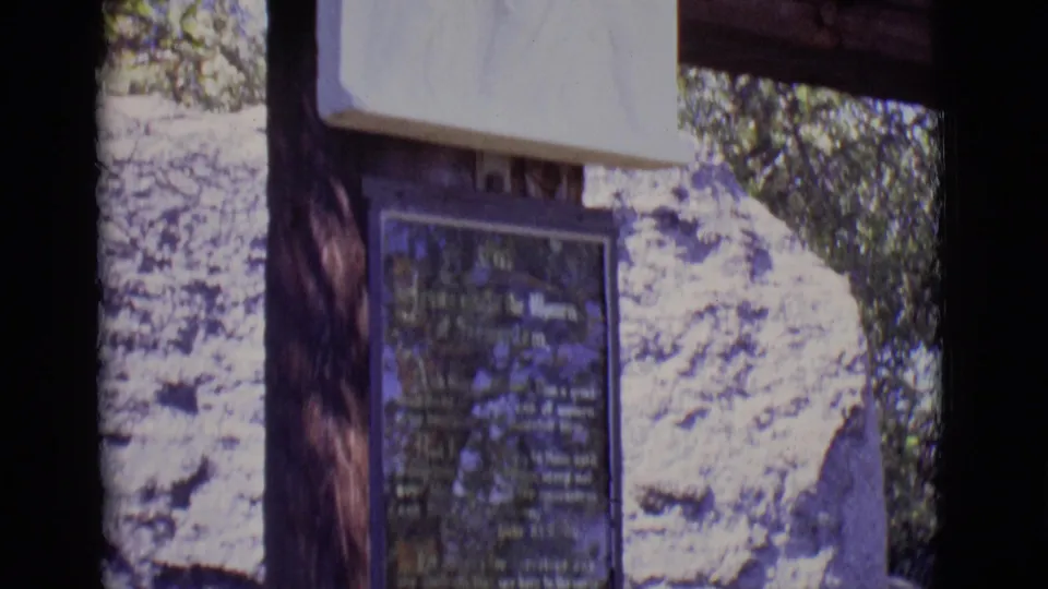 1965 San Luis, Colorado frame showing the lower plaque beneath a white Stations of the Cross relief on an outdoor wooden structure.