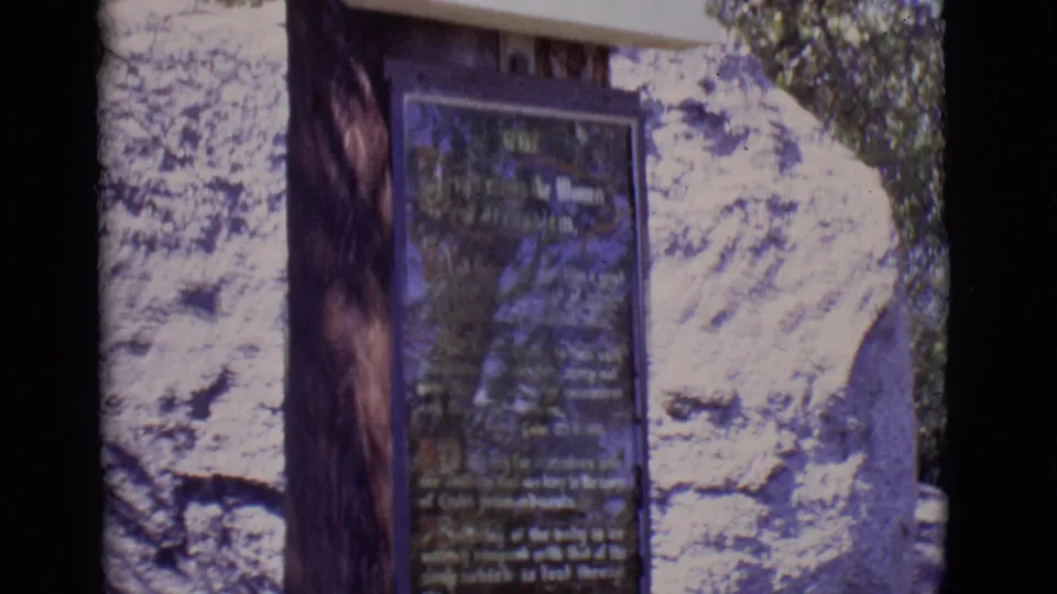 The final frame centers on the explanatory plaque beneath the religious relief, with stone, wood, and foliage surrounding it.