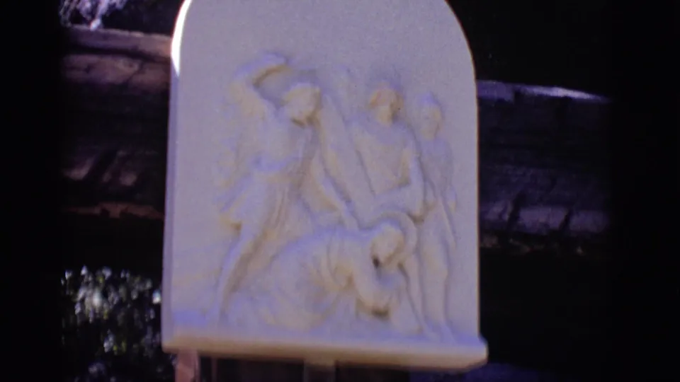 The frame centers the carved Ninth Station relief in San Luis, Colorado, with pale stone figures and the darker explanatory plaque partly visible underneath.