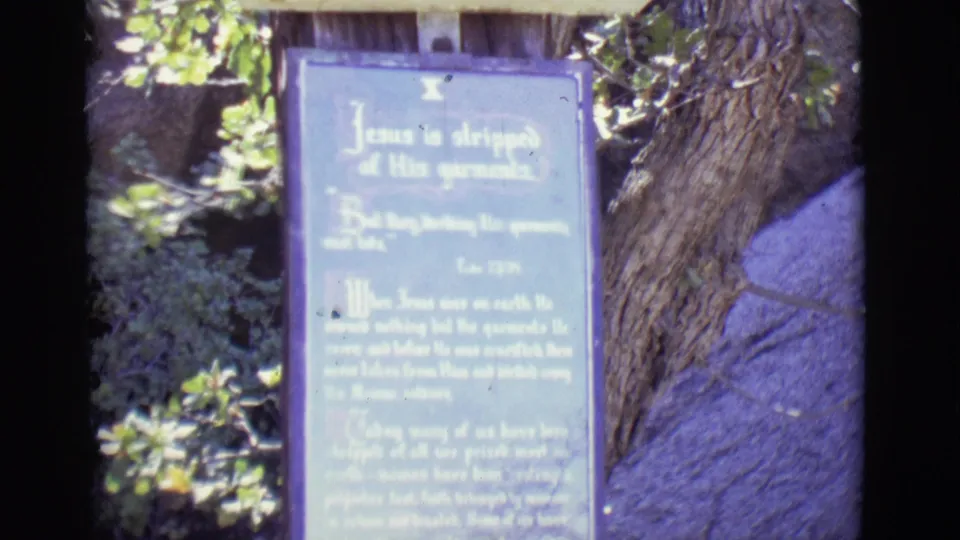 1965 outdoor frame in San Luis, Colorado showing a blue Stations plaque about Jesus stripped of garments near a tree trunk and foliage.