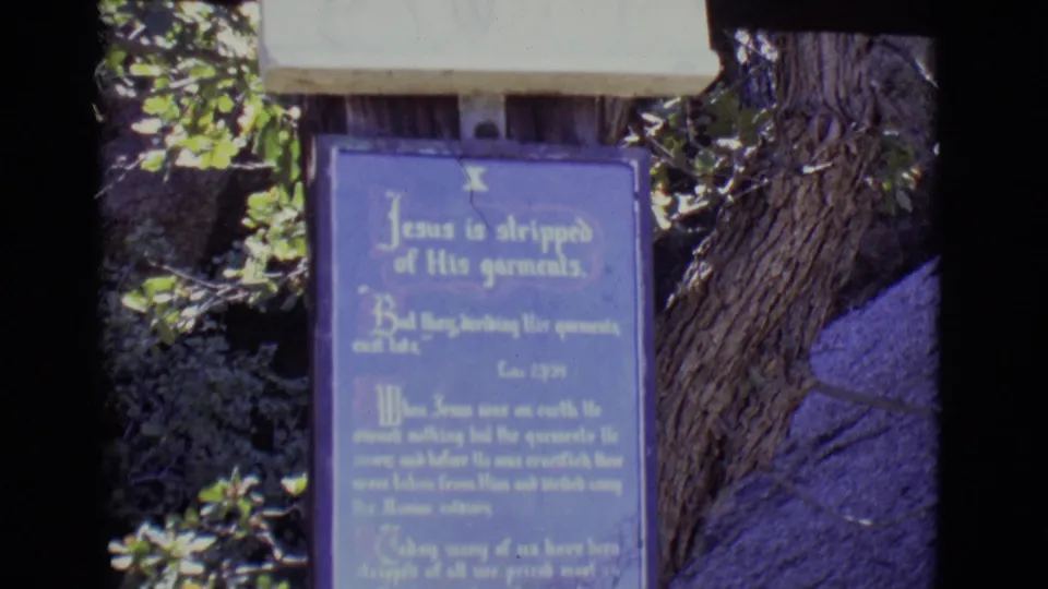 Tight 8mm view of the tenth Stations of the Cross plaque, mounted outdoors below a pale relief base with trees and rock nearby.