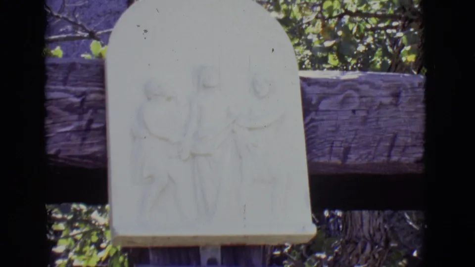 1965 San Luis, Colorado 8mm view of a pale carved relief showing religious figures near a wooden rail and trees.
