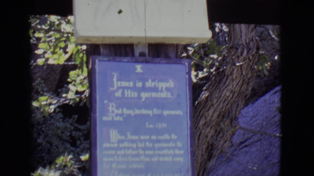 Blue religious plaque hanging outdoors beside tree trunks and foliage.