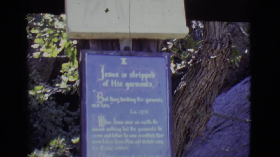 1965 San Luis, Colorado 8mm frame showing a blue plaque about Jesus being stripped of garments along an outdoor religious path.