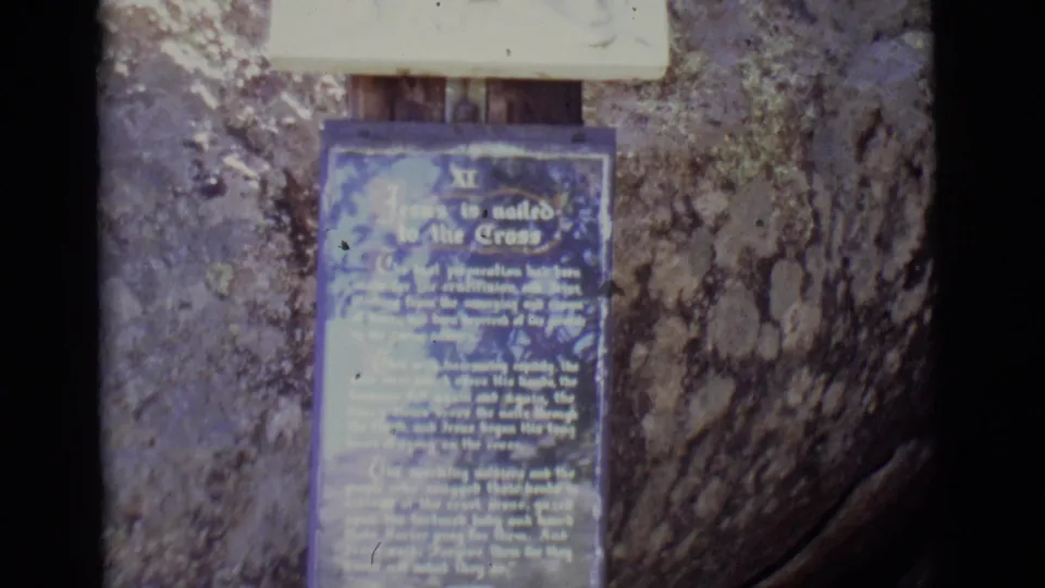 1960s 8mm frame of a blue devotional plaque with white text about Jesus being nailed to the cross, mounted outdoors near bark and wood.