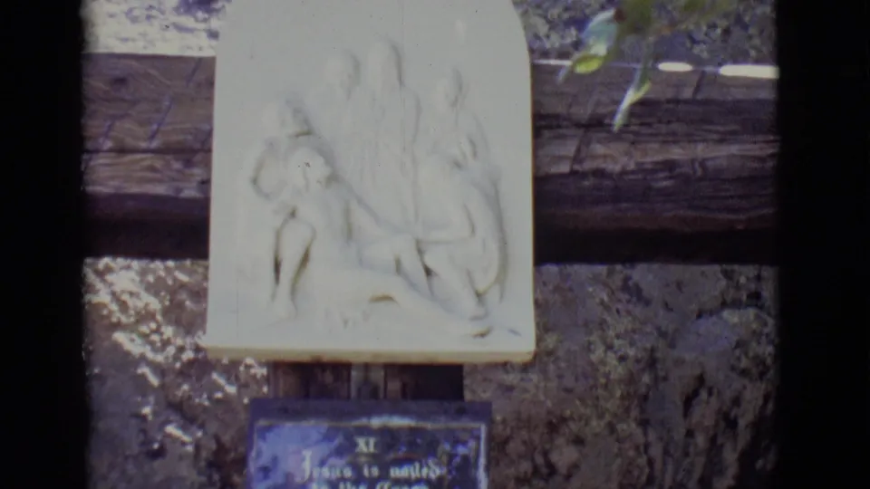 A pale relief sculpture of a religious scene appears above a blue plaque on an outdoor path in San Luis, Colorado, filmed on 8mm.