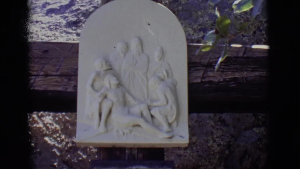 1965 San Luis, Colorado 8mm frame showing a white relief sculpture above a weathered stone wall and wooden cross support.