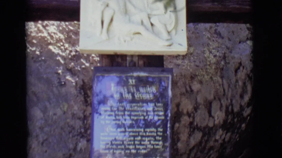 1965 home-movie frame showing the lower relief edge and a blue explanatory plaque, with text softened by focus and glare.