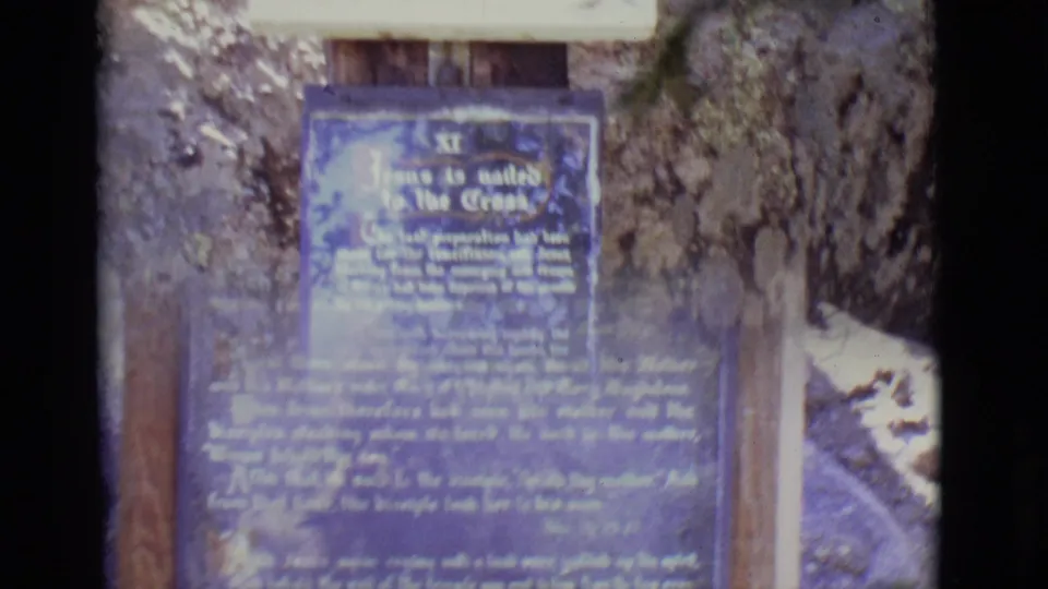 1965 San Luis, Colorado 8mm frame showing a close view of a blue devotional sign on wood posts beside rough stone and rocks.