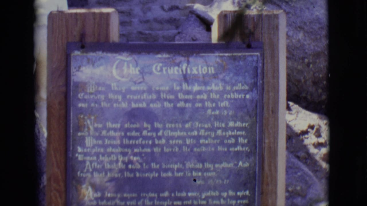 1965 San Luis, Colorado frame with a blue sign headed The Crucifixion, mounted outdoors on wooden posts before rocks and stonework.