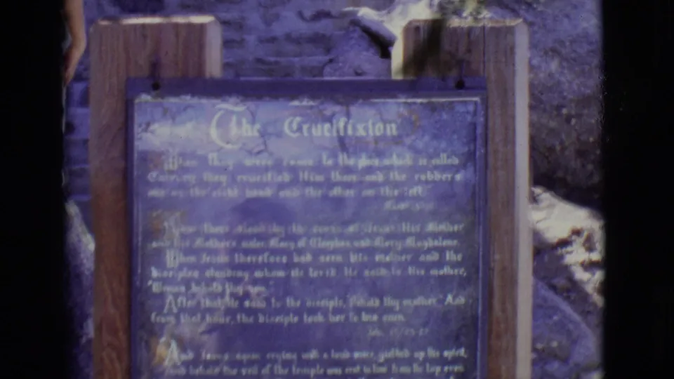Close 8mm view of a blue outdoor crucifixion narrative sign in San Luis, Colorado, with wood posts and rocks visible around it.