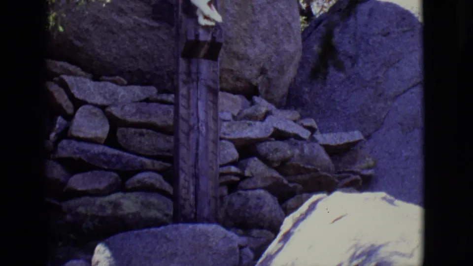 1965 San Luis, Colorado 8mm frame showing a narrow wooden post or marker among rocks and stacked stone near the religious display.