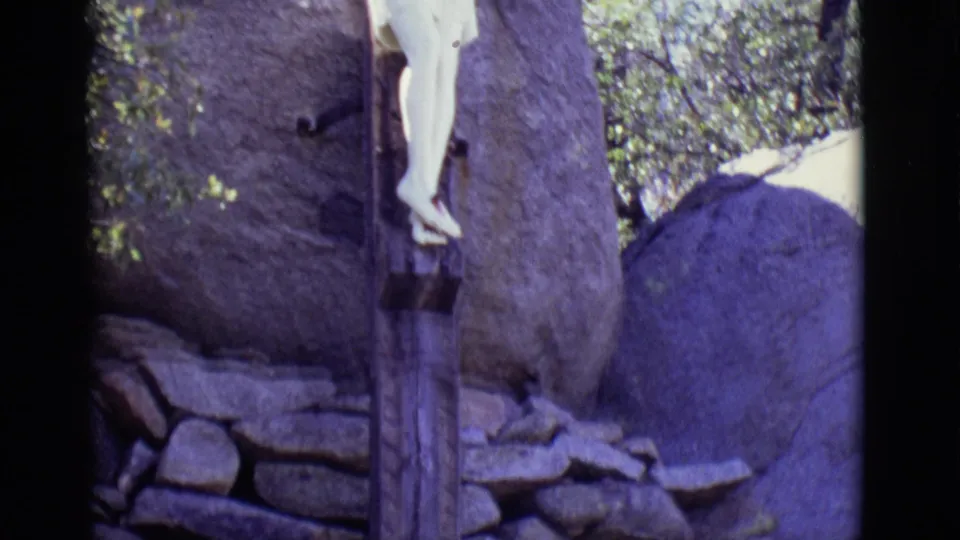 1965 San Luis, Colorado 8mm frame showing the lower part of a white crucifix statue near boulders, stonework, trees, and dark film borders.