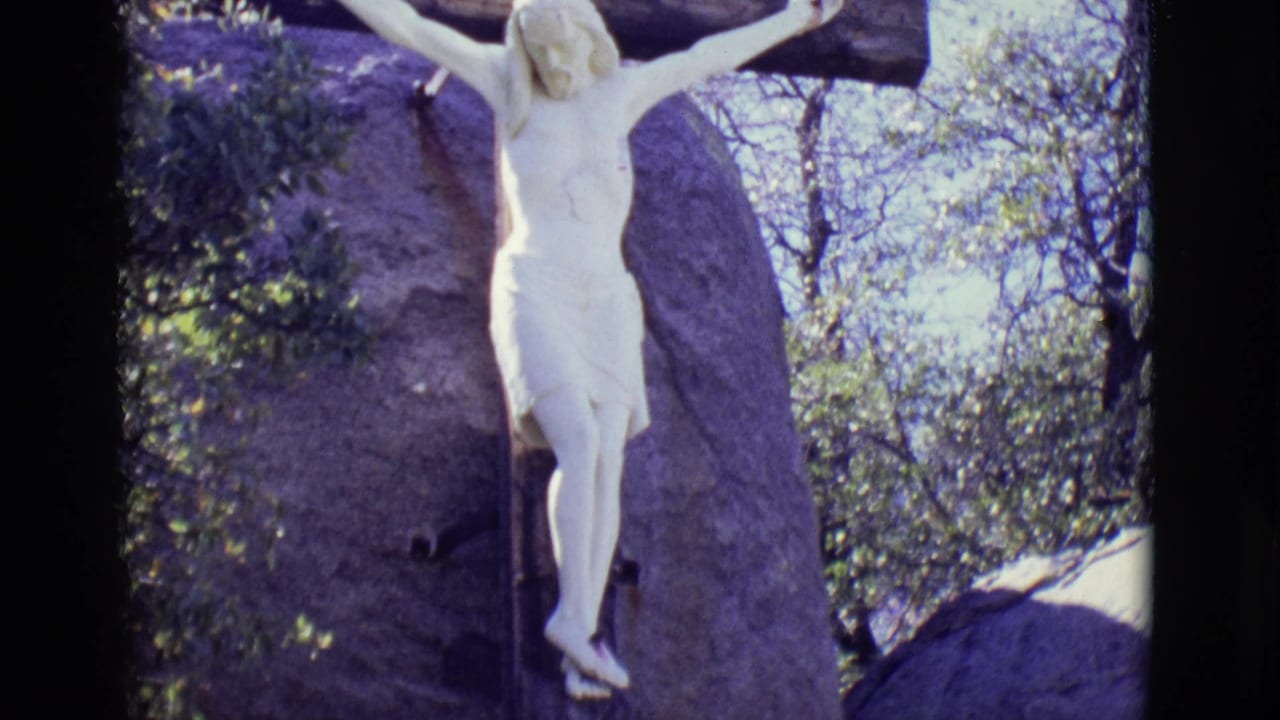 White crucifix statue mounted on a dark wooden cross at the San Luis, Colorado pilgrimage site, with boulders and leafy trees in the background.