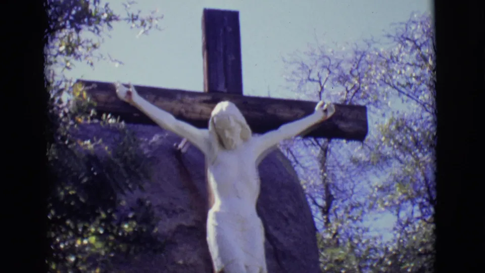 Close 8mm view of the upper crucifix in San Luis, Colorado, showing the outstretched arms, wooden crossbeam, rock backdrop, trees, and pale sky.