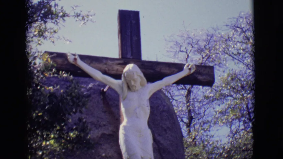 Final 1965 home-movie frame of the white crucifix at San Luis, Colorado, framed by a wooden cross, boulders, trees, bright sky, and rounded film edges.