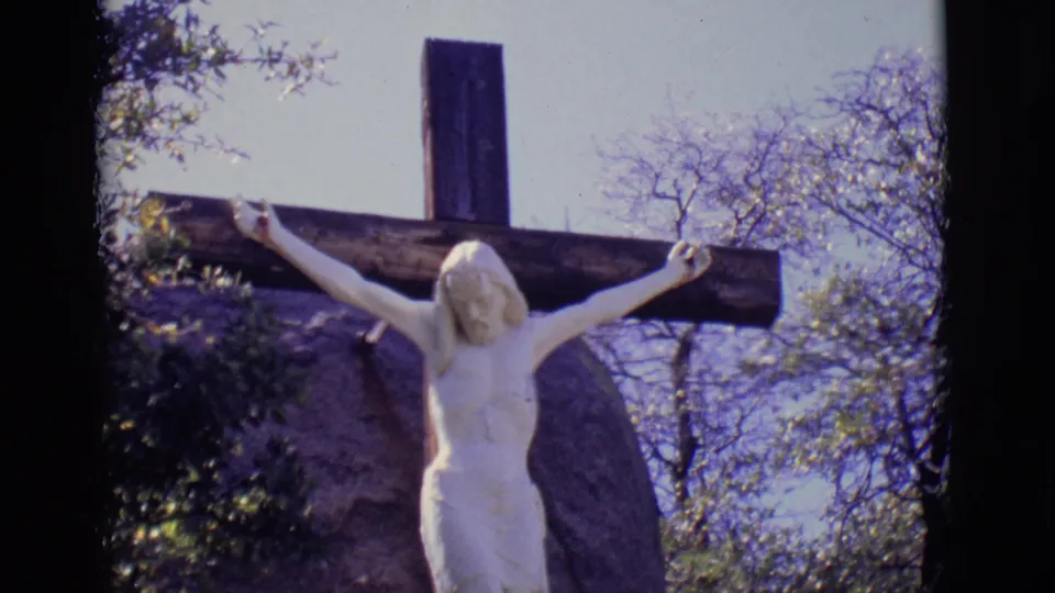 1965 8mm frame from San Luis, Colorado, showing a white Jesus crucifix statue on a dark cross with trees and sky behind it.