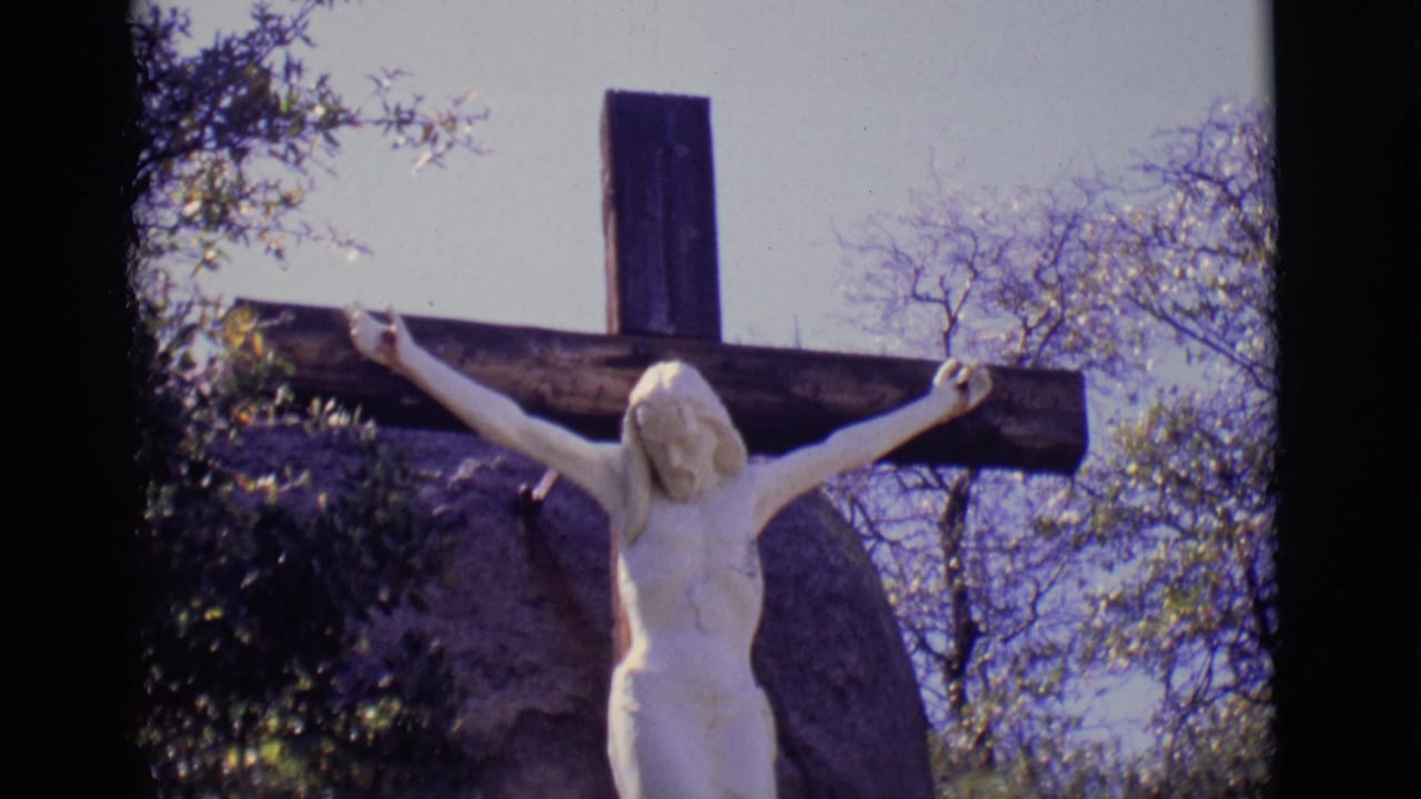 White crucified Jesus statue outdoors in a 1965 San Luis, Colorado home-movie frame, with a wooden cross and sparse trees visible.
