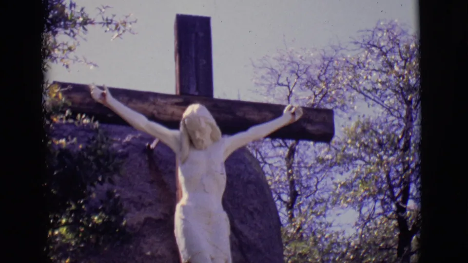 1965 8mm view of an outdoor crucifix figure in San Luis, Colorado, with the cross rising above trees and a stone or rock form.