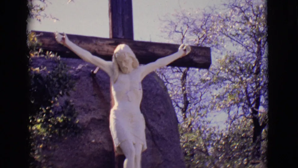 Final 1965 home-movie frame of the San Luis, Colorado crucifix statue, with the white figure, dark cross, trees, and pale sky visible.