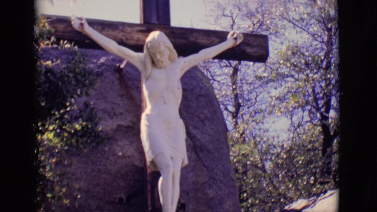 White crucifixion statue on a wooden cross beside a large rock and trees.