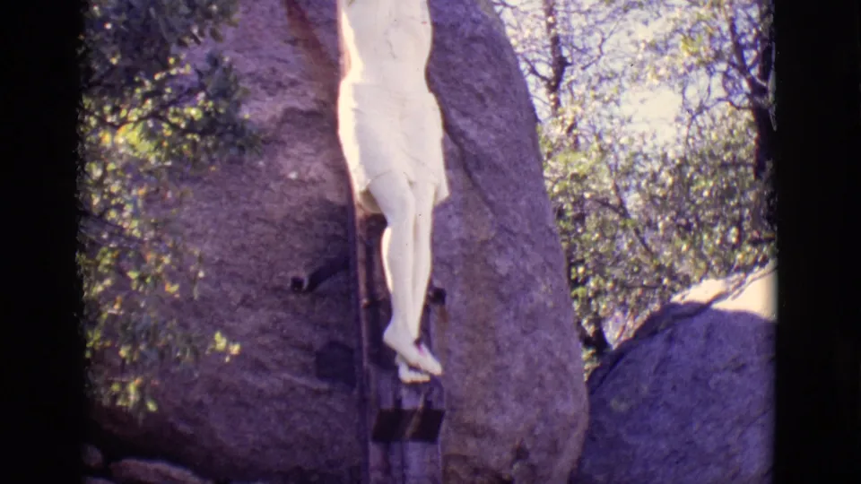 Tighter 8mm frame showing the lower body of the crucifixion statue, the vertical wooden post, and the adjacent rock surface.