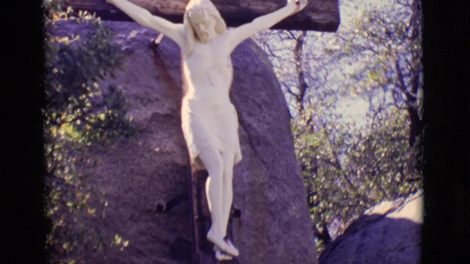 San Luis, Colorado home-movie frame showing the crucifixion statue centered against a large rock and leafy outdoor background.