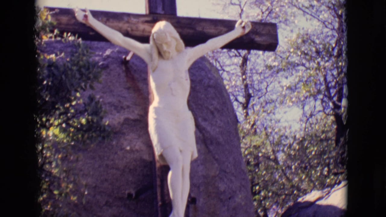 White Jesus crucifix statue on a dark wooden cross beside rock and trees.