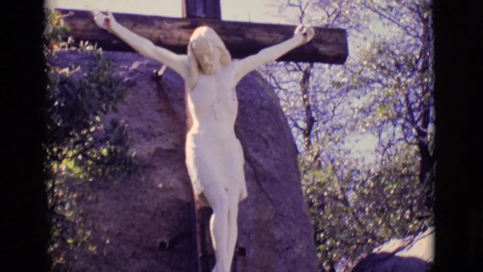 1965 San Luis, Colorado 8mm frame showing a white crucifix statue against a wooden cross, rock, shrubs, and pale sky.