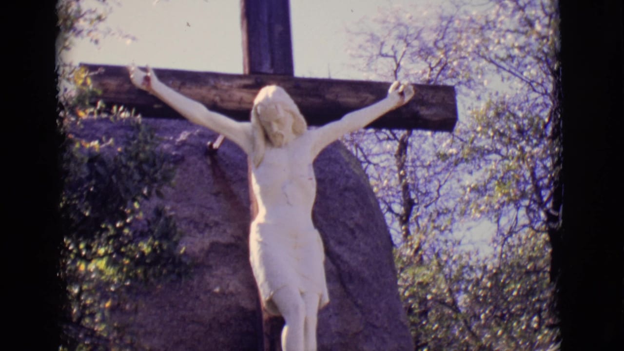 1965 San Luis, Colorado frame with the crucifix statue centered against a dark cross, rock face, branches, and sky.