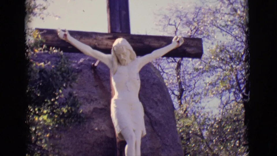 1965 San Luis, Colorado frame with the crucifix statue centered against a dark cross, rock face, branches, and sky.