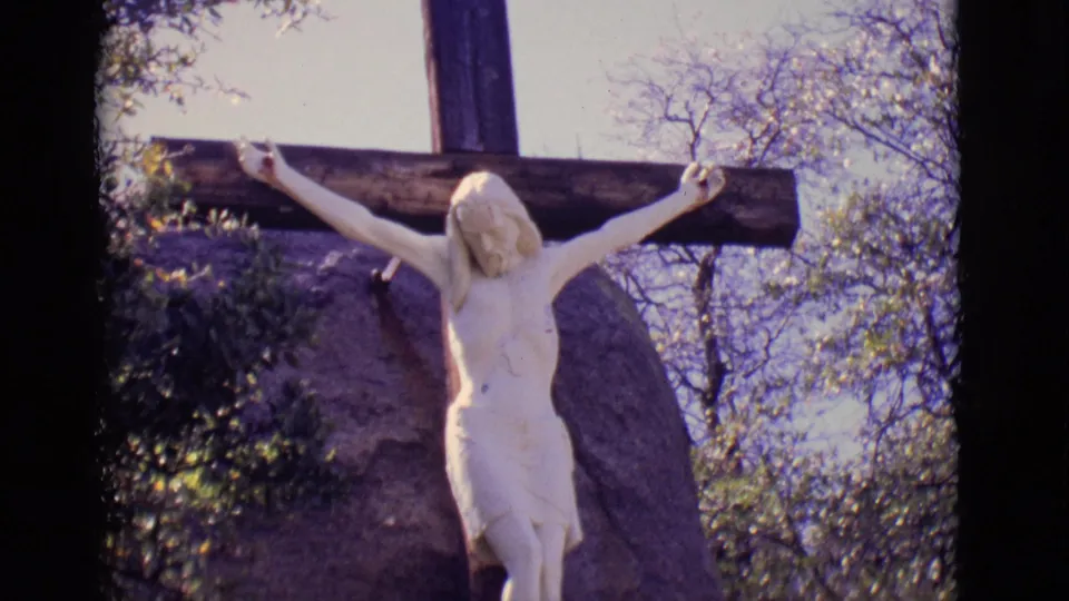 1965 San Luis, Colorado 8mm frame showing the crucifix statue closer in view, with rock, shrubs, tree branches, and sky behind.