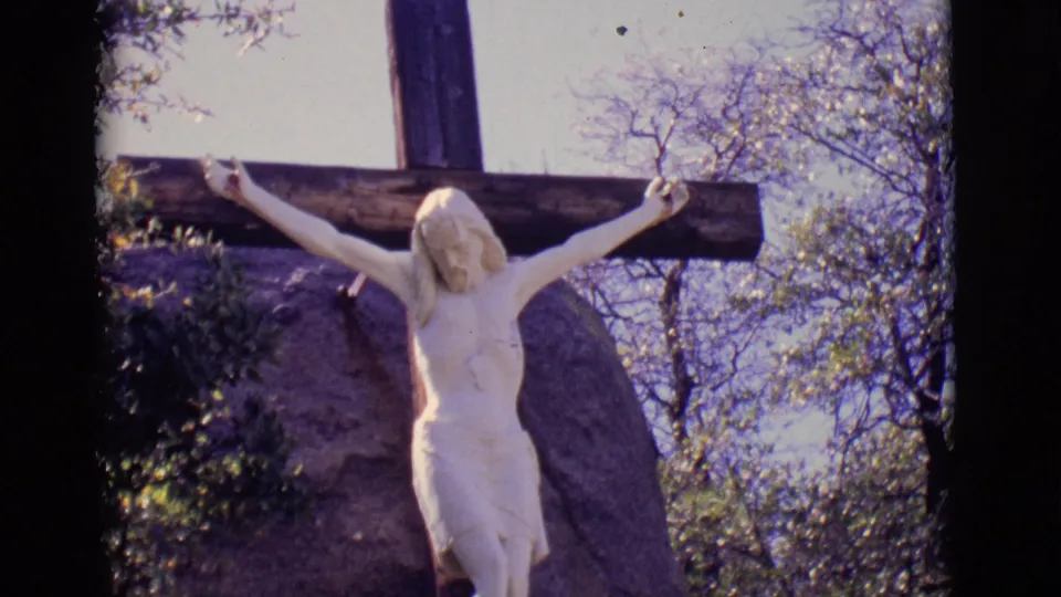 1965 San Luis, Colorado frame of a white Jesus crucifix statue on a wooden cross, surrounded by rock, shrubs, and wintery branches.