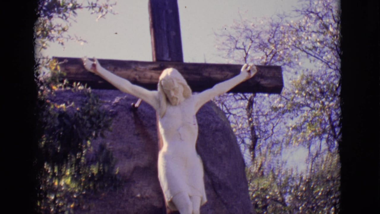 1965 San Luis, Colorado 8mm frame showing a white crucified Jesus statue on a dark wooden cross beside trees and a large boulder.