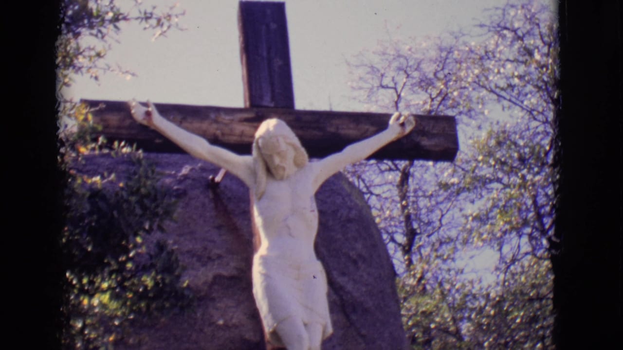 1965 San Luis, Colorado 8mm view of a white crucifix statue mounted on a wooden cross with trees, sky, and stone behind it.
