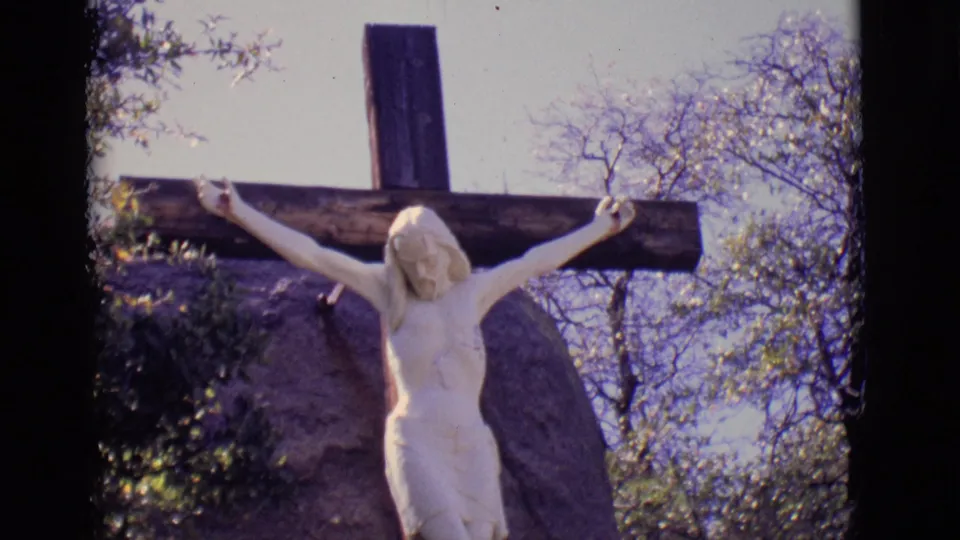 1965 San Luis, Colorado home-movie frame with the crucified Jesus statue slightly lower in view, framed by rock, shrubs, and branches.