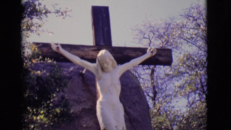 1965 San Luis, Colorado 8mm frame of a white crucified Jesus statue fixed to a wooden cross outdoors near a large boulder and trees.
