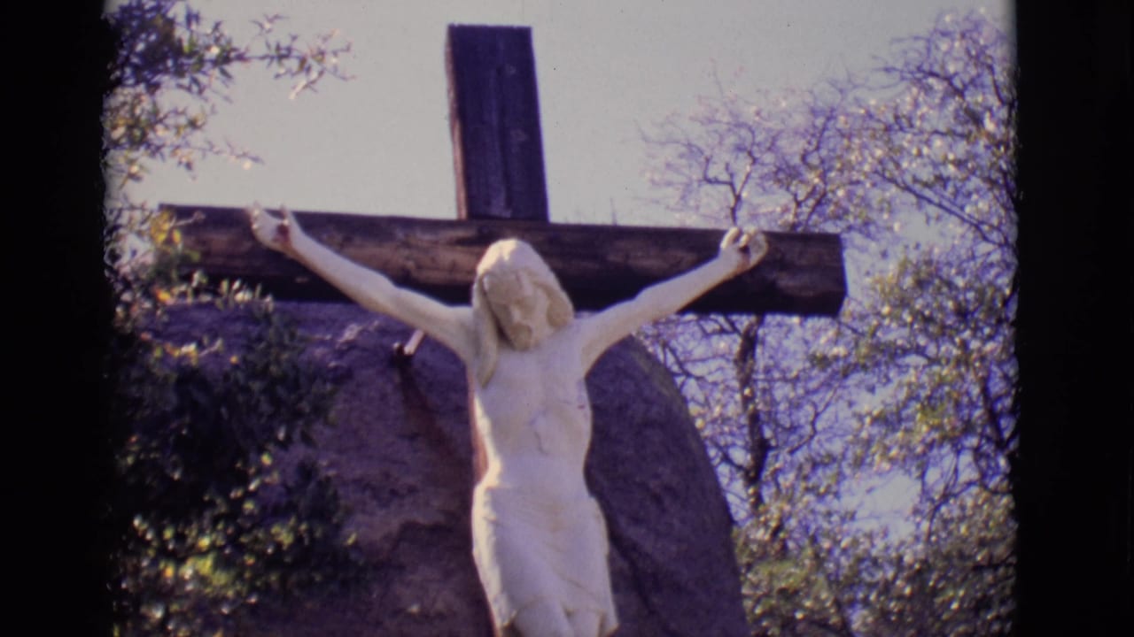1965 San Luis, Colorado 8mm frame showing a white Jesus statue on a dark wooden cross with rocks, shrubs, and trees behind it.