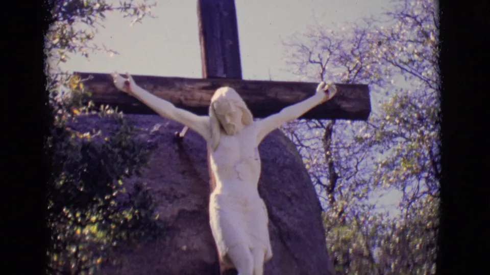 1965 San Luis, Colorado 8mm frame with a closer view of the white Jesus statue on the cross, stone setting, shrubs, and bright sky.