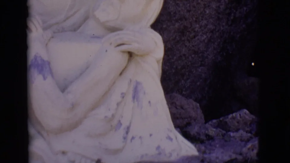 1965 8mm frame from San Luis, Colorado showing a close view of a pale stone Mary figure cradling Jesus near a dark rock face.