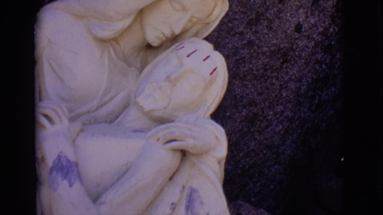 1965 San Luis, Colorado frame showing a pale stone religious statue of Mary holding Jesus, with red forehead marks and dark stone behind it.