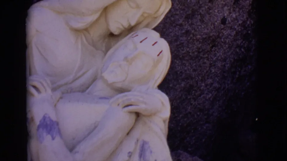 1965 San Luis, Colorado frame showing a pale stone religious statue of Mary holding Jesus, with red forehead marks and dark stone behind it.