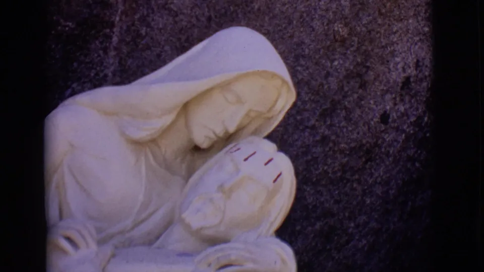 1965 8mm frame from San Luis, Colorado showing Mary leaning over Jesus in a pale stone sculpture, closely framed against dark rock.