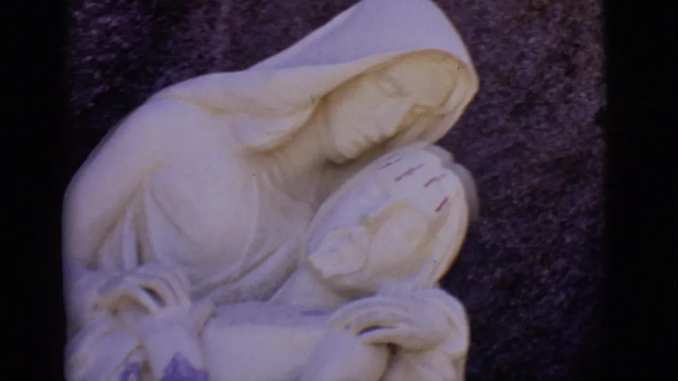 Final 1965 San Luis, Colorado frame showing the pale stone Mary and Jesus statue with carved hands, drapery, and dark rock background.