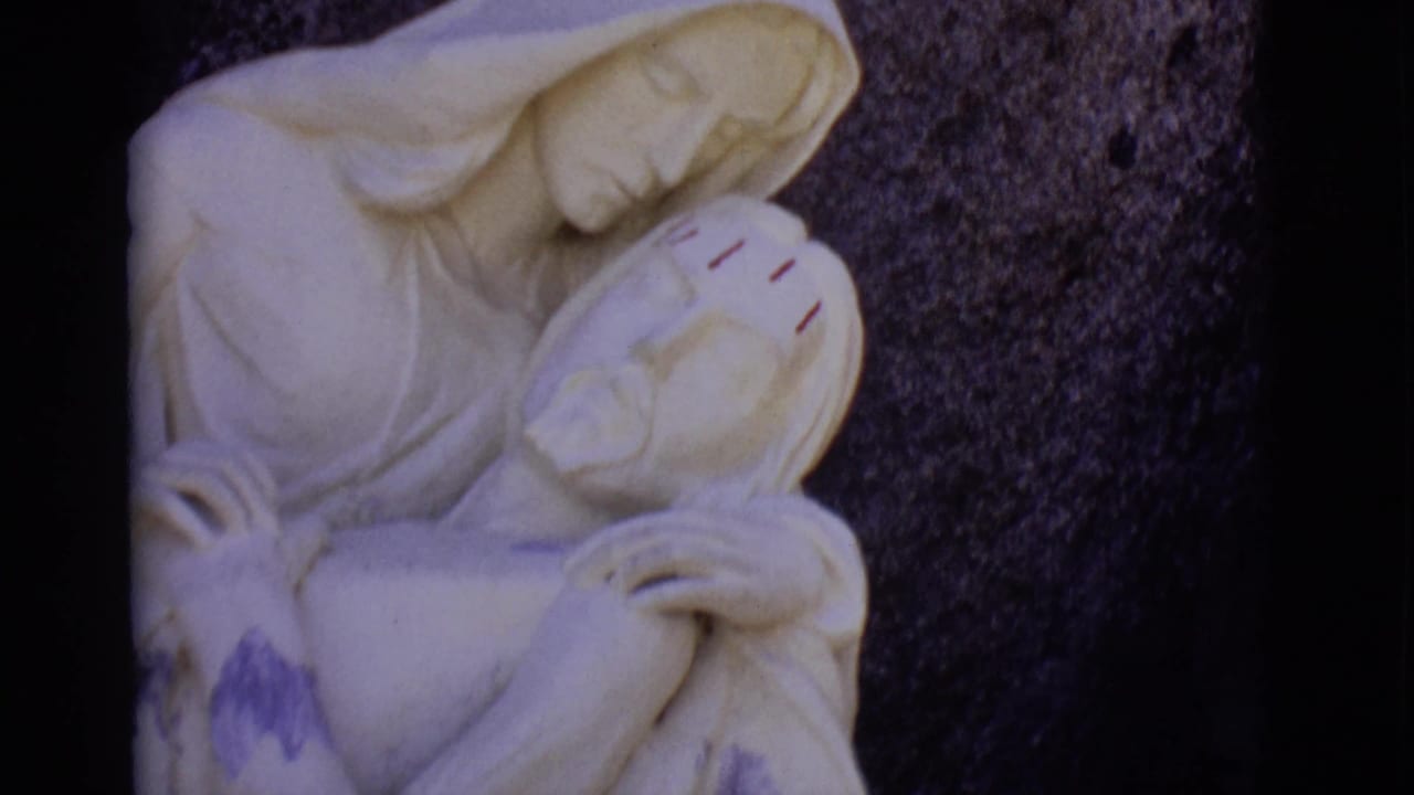 White devotional statue showing Mary holding Jesus against a dark stone background.