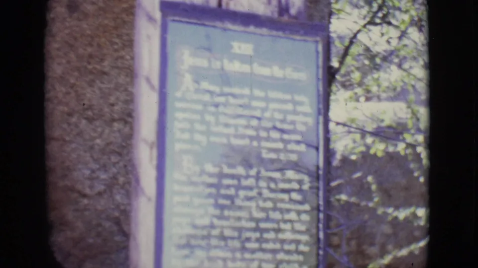 1965 San Luis, Colorado frame showing a blue station-of-the-cross plaque beside stonework and sunlit foliage.