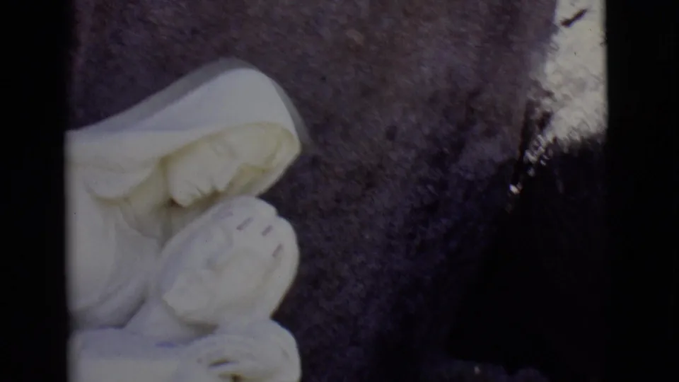 1965 San Luis, Colorado 8mm frame with a white marble Virgin Mary figure cradling Jesus against a dark stone backdrop.