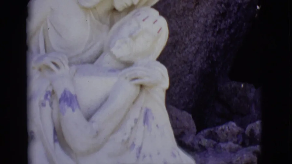 1965 San Luis, Colorado 8mm frame showing a pale Pieta sculpture with Mary cradling Jesus before a dark rock surface.