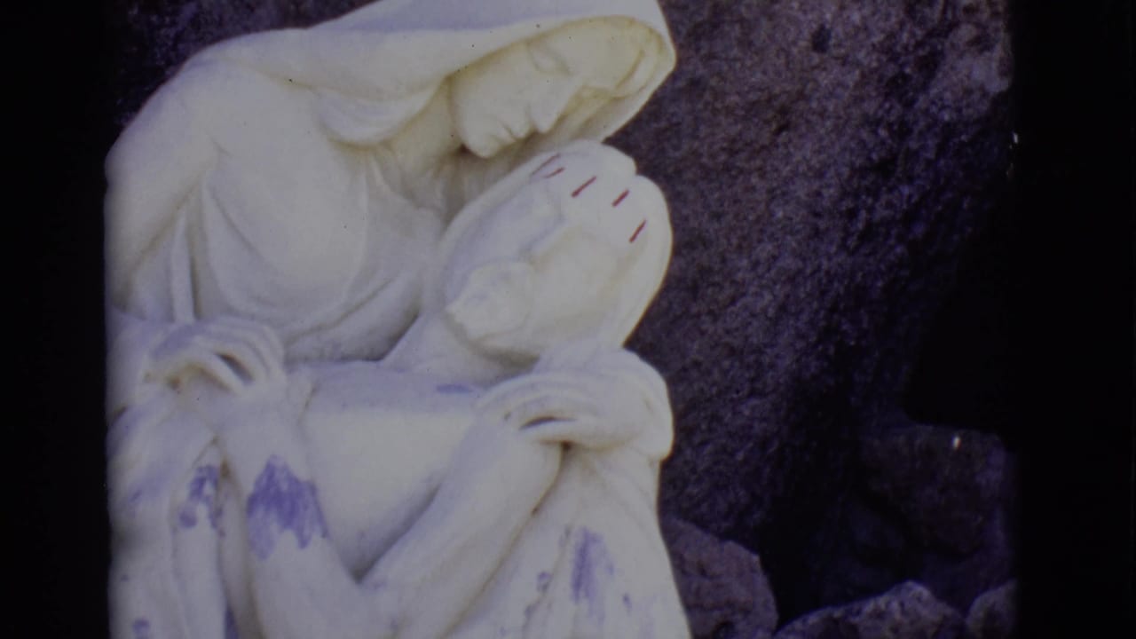 San Luis, Colorado home-movie frame of the Pieta sculpture, showing Mary bowed over Jesus with dark stone behind them.