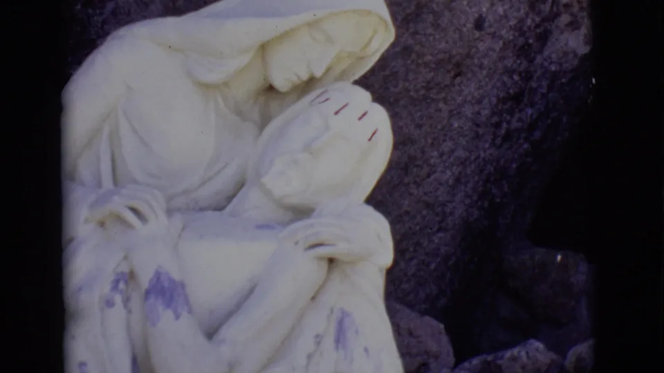 San Luis, Colorado home-movie frame of the Pieta sculpture, showing Mary bowed over Jesus with dark stone behind them.